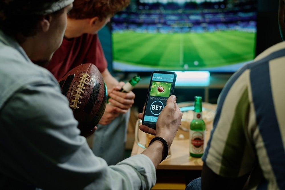 An overhead view of a sports betting setup, featuring a smartphone displaying betting odds, a notepad with handwritten notes, and a tabletop filled with sports paraphernalia, including a football, basketball, and racing information, symbolizing the excitement of sports wagering.