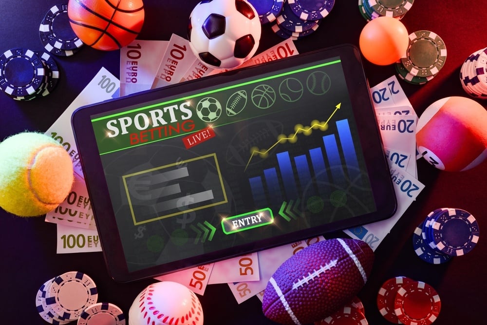 An overhead view of a sports betting setup, featuring a smartphone displaying betting odds, a notepad with handwritten notes, and a tabletop filled with sports paraphernalia, including a football, basketball, and racing information, symbolizing the excitement of sports wagering.